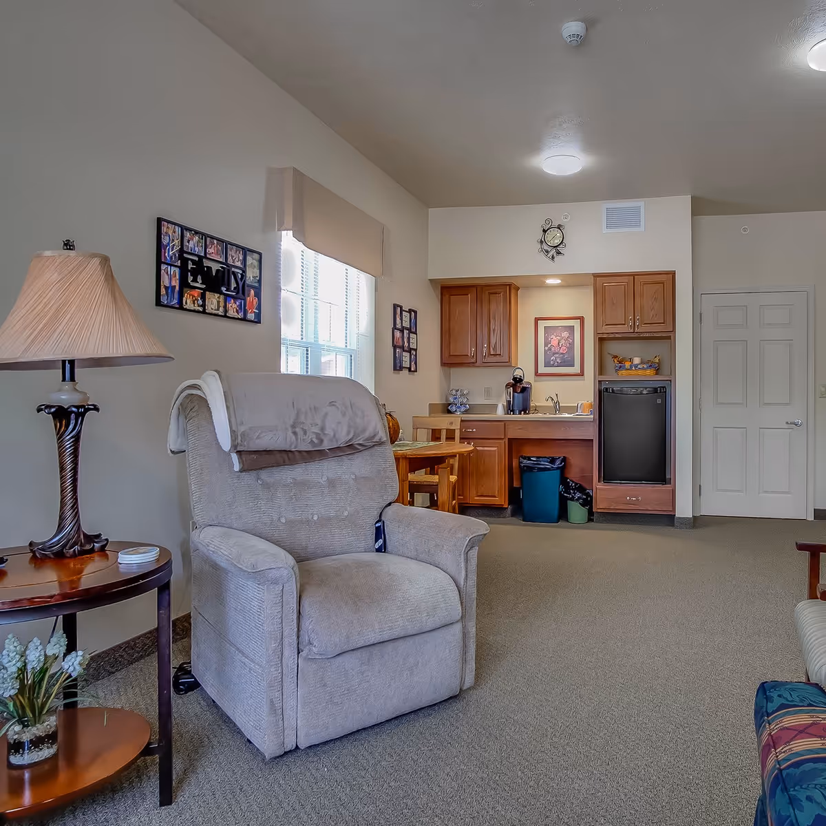 A comfortable living room area with a beige recliner and side table lamp in the foreground and a small kitchenette with cabinets and a table in the background.