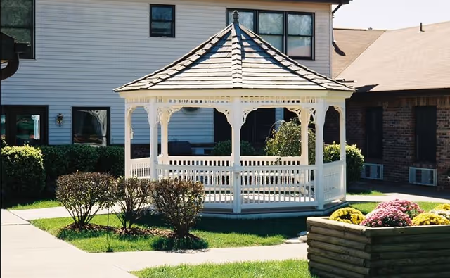 A white wooden gazebo with a shingled roof situated on a grassy area surrounded by bushes and flowers, with residential buildings in the background.