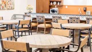 Bright dining area with round wooden tables and patterned chairs in front of a counter and cabinets.
