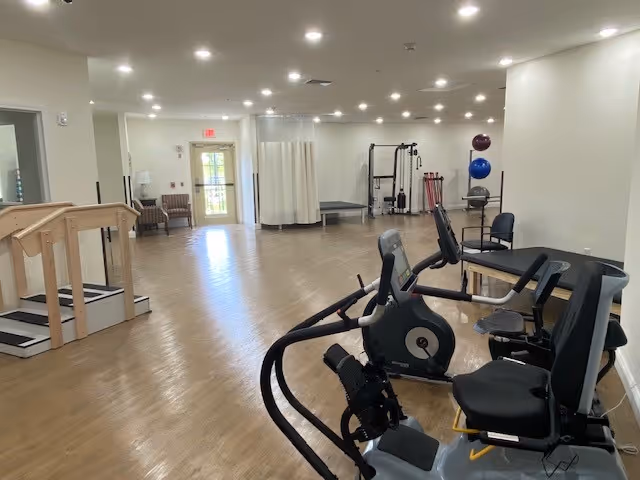 A spacious rehabilitation room with exercise equipment including stationary bikes and parallel bars. The room has wooden flooring, white walls, and bright ceiling lights. There are chairs and a curtained area near the back, with a door leading outside.