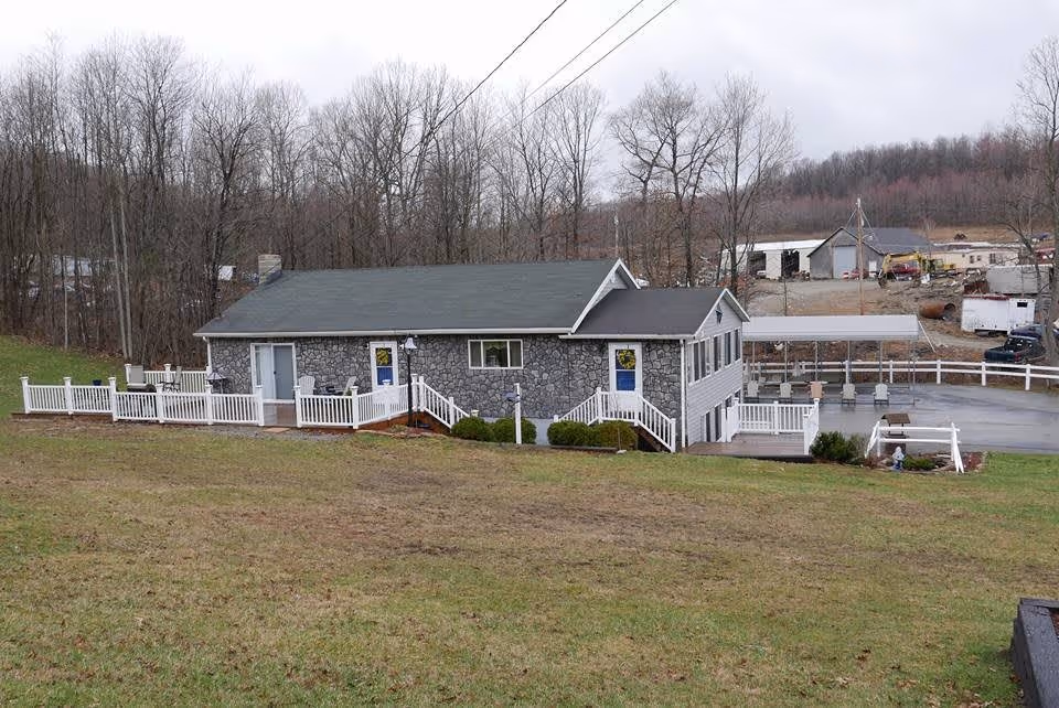 A single-story stone and siding building with a gray roof, white railings, and multiple entrances, situated on a grassy slope with leafless trees in the background and some industrial buildings further behind.
