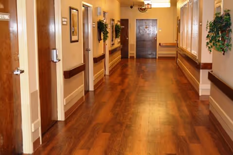Well-lit interior hallway with polished wood floors, doors and handrails along the sides, wall plants, and double doors at the far end.