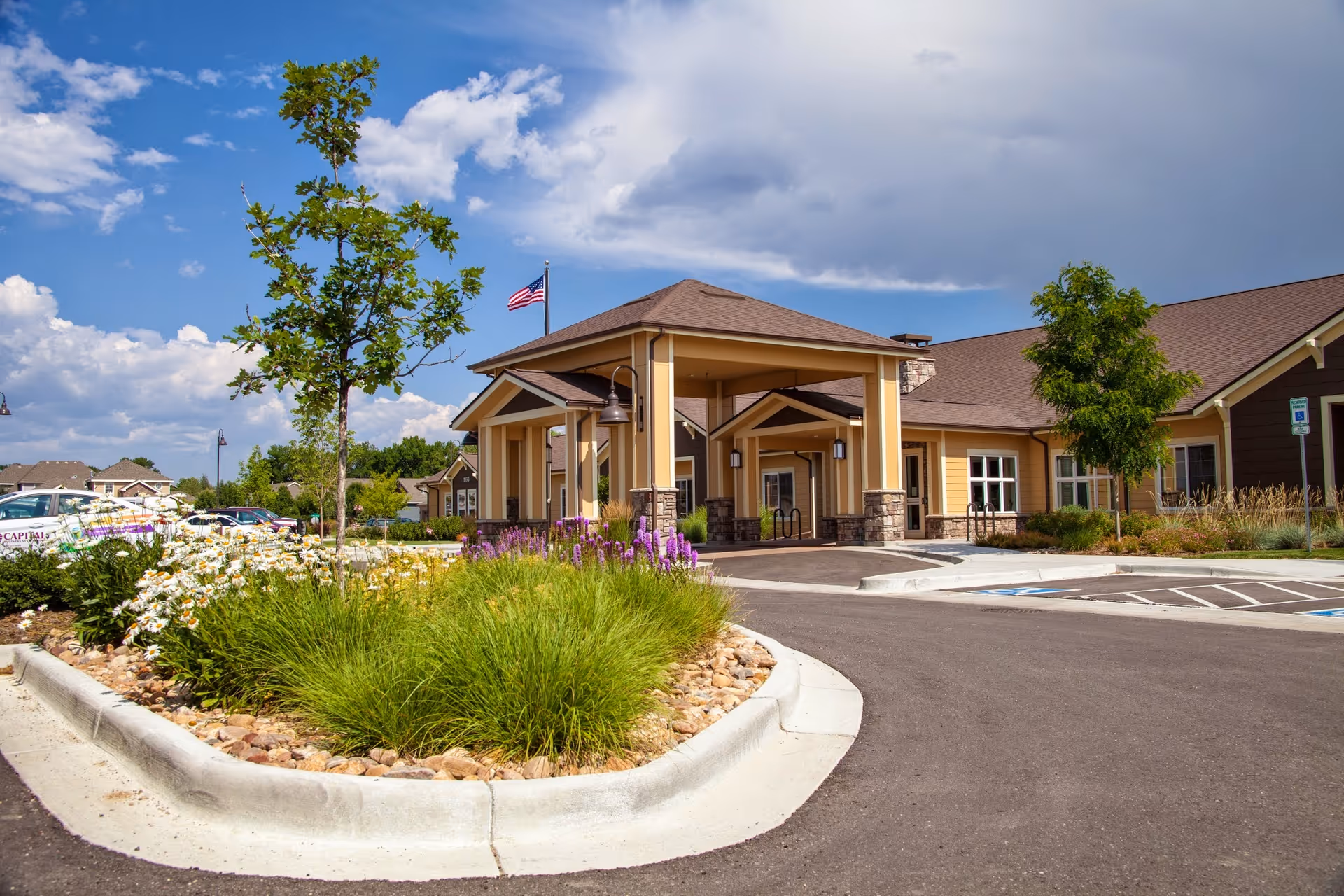 Exterior view of Seven Lakes Memory Care facility showing the entrance with a covered drop-off area, landscaped garden with flowers and greenery, an American flag on a pole, and a clear blue sky with some clouds.