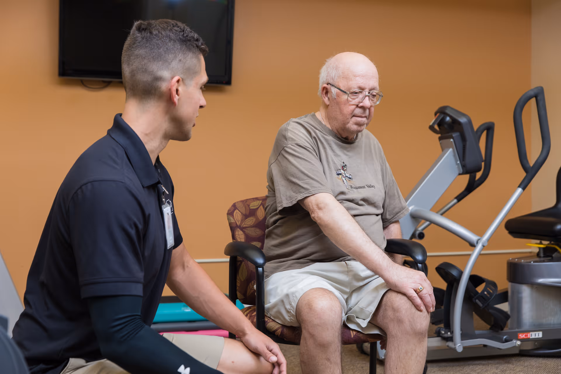 An elderly man sitting on a chair in a fitness room, touching his knee, while a younger man in a black polo shirt kneels beside him, appearing to assist or talk to him. Exercise equipment is visible in the background.
