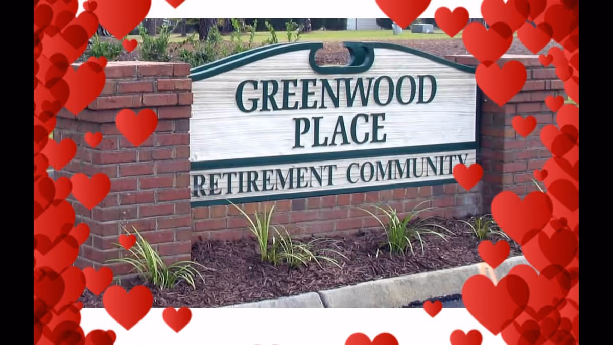 A brick sign at the entrance of Greenwood Place Retirement Community with landscaping around it. The image is decorated with red heart graphics around the edges.