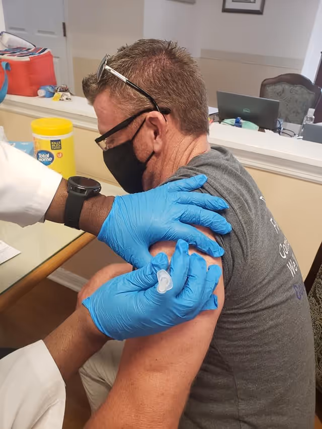 A man wearing a black face mask and glasses on his head is receiving a vaccine injection in his upper arm from a healthcare worker wearing blue gloves. The setting appears to be an indoor office or clinic area with a desk, laptop, and cleaning supplies in the background.