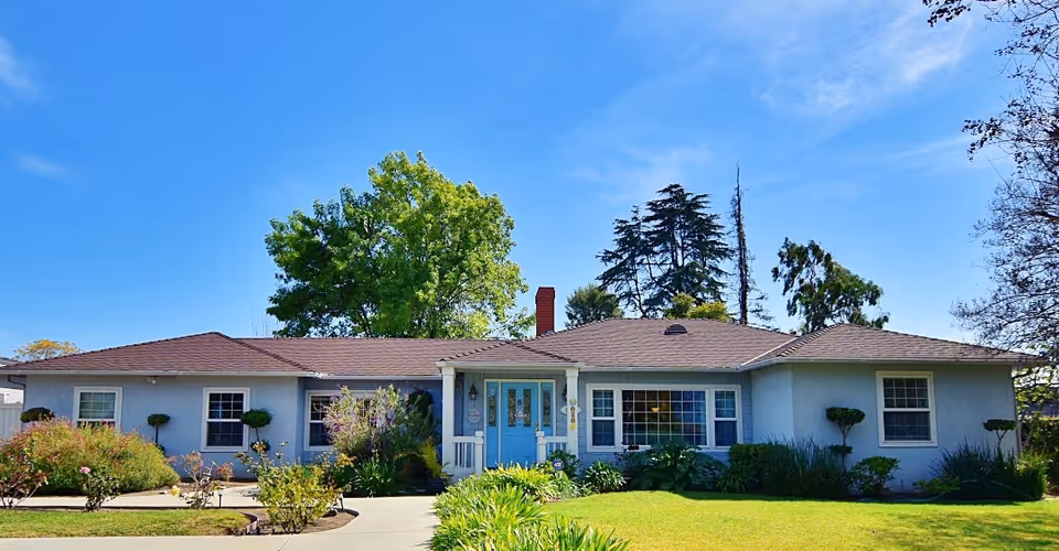Single-story ranch-style residence with a blue front door, landscaped lawn and trees under a clear blue sky.