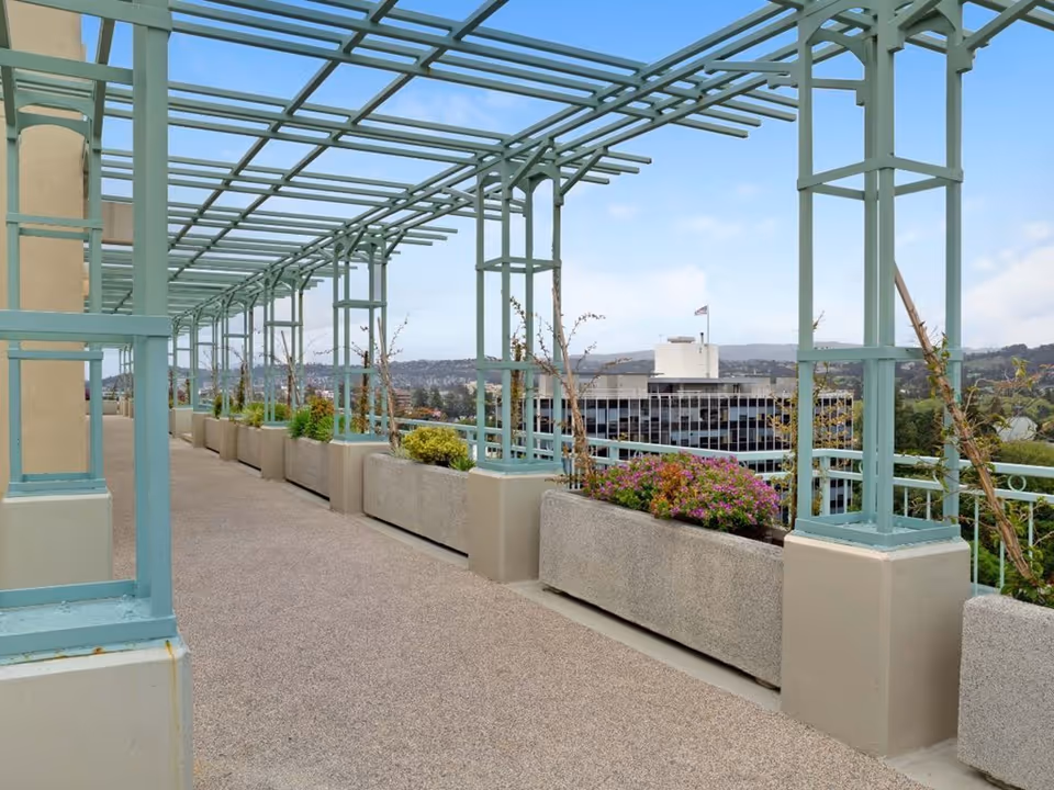 A rooftop terrace walkway with teal pergola structures and concrete planter boxes overlooking surrounding buildings.