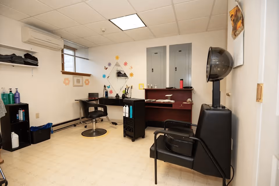 Interior of a small hair salon room with a salon chair in front of a mirror decorated with colorful flower stickers. There are shelves with hair products, a hair dryer chair, and a desk with various items. The room has a window with blinds, an air conditioning unit, and a tiled floor.