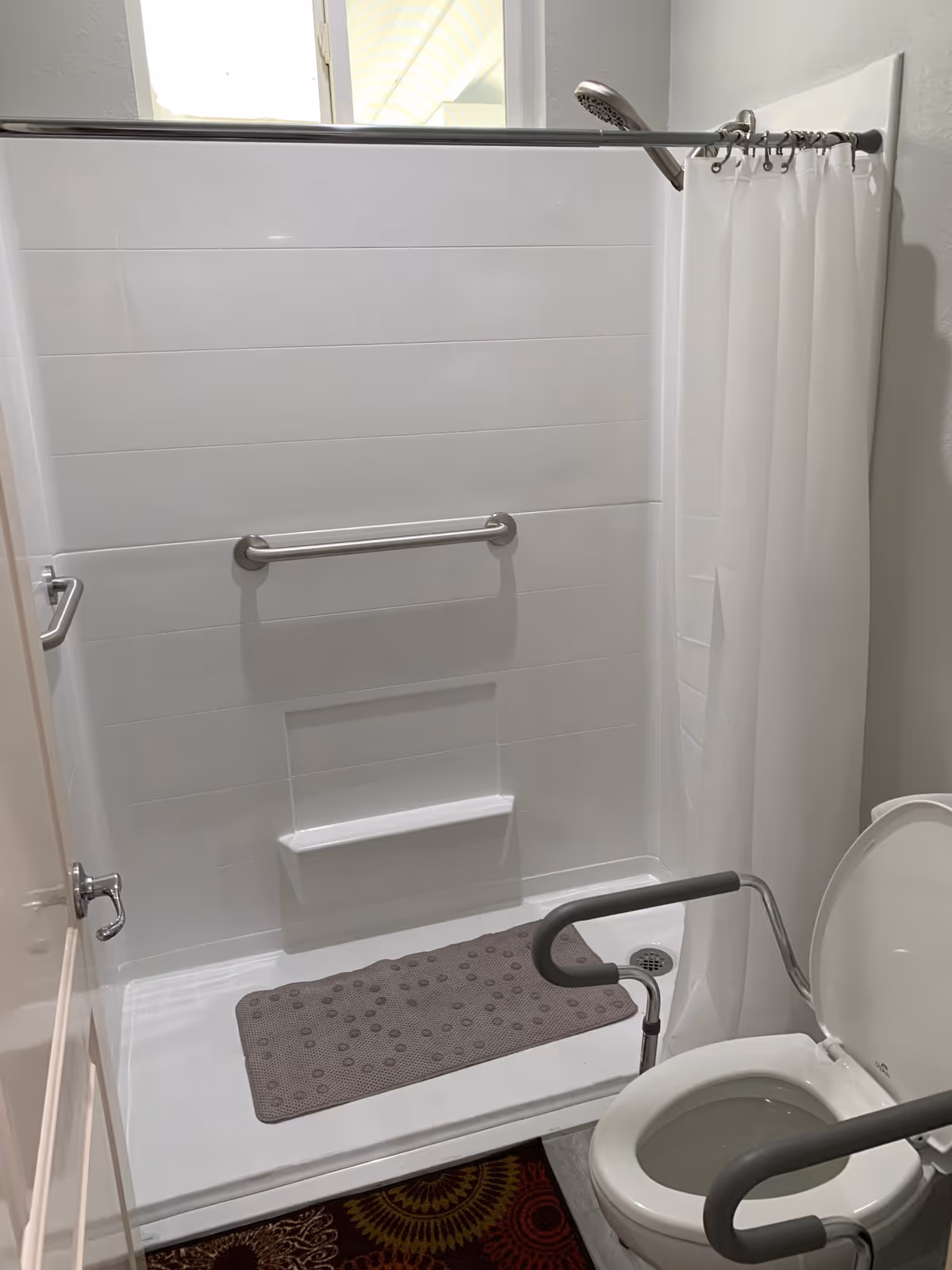 A bathroom with a white tiled shower area featuring a grab bar and a small built-in shelf. There is a gray non-slip mat on the shower floor and a white shower curtain partially drawn. In the foreground, there is a toilet with gray safety rails on both sides. The bathroom appears clean and designed for accessibility.