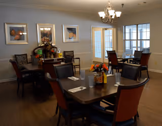 A dining room in an assisted living facility with several wooden tables and chairs arranged neatly. Each table has place settings with napkins and glasses, and some have small flower arrangements. The room has light-colored walls adorned with framed artwork and a chandelier hanging from the ceiling. Large windows and glass doors allow natural light into the space.