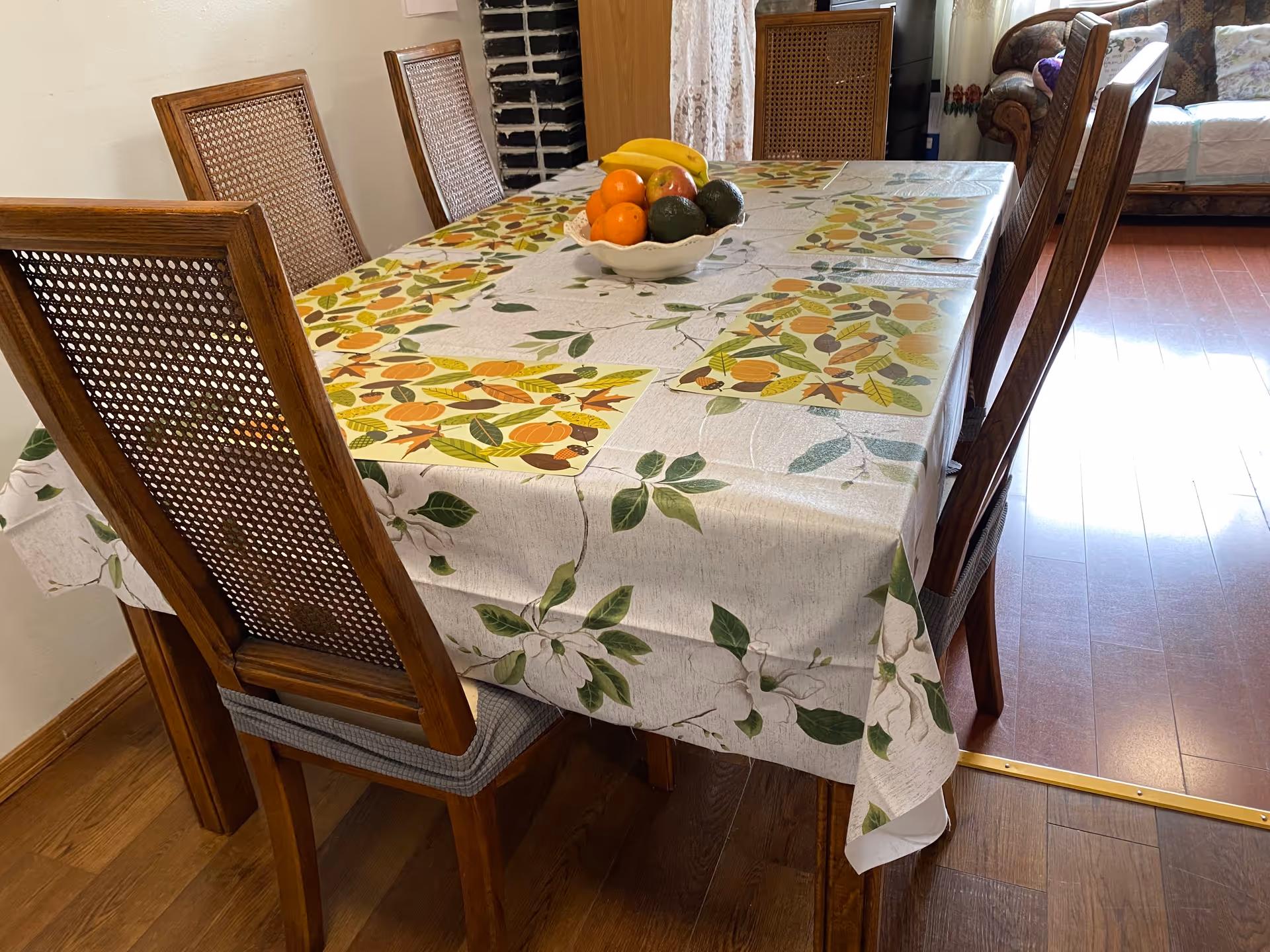 A dining table covered with a floral tablecloth and four placemats featuring autumn leaves. On the table, there is a white bowl filled with bananas, oranges, an apple, and avocados. The table is surrounded by wooden chairs with woven backs. In the background, there is a couch and a wooden cabinet.