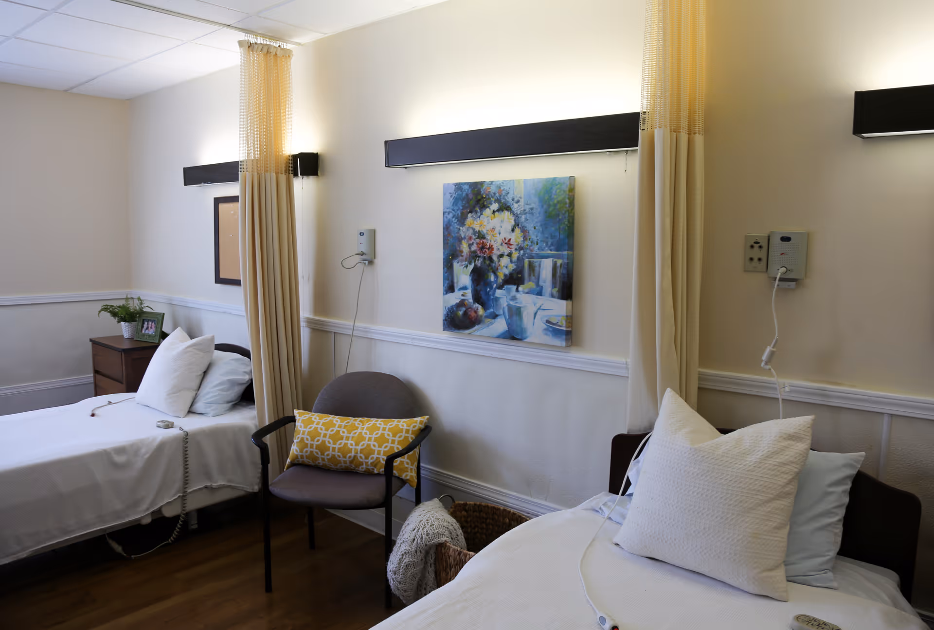 Shared patient bedroom with two beds, a chair, bedside table, wall art and privacy curtains.
