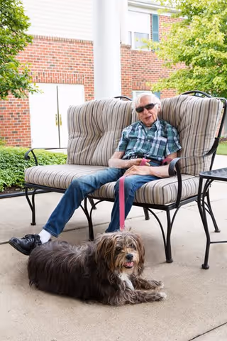 An elderly man wearing sunglasses and a plaid shirt sits on a striped cushioned outdoor bench holding a pink leash attached to a small, fluffy dog lying on the ground in front of him. They are outside near a brick building with greenery around.