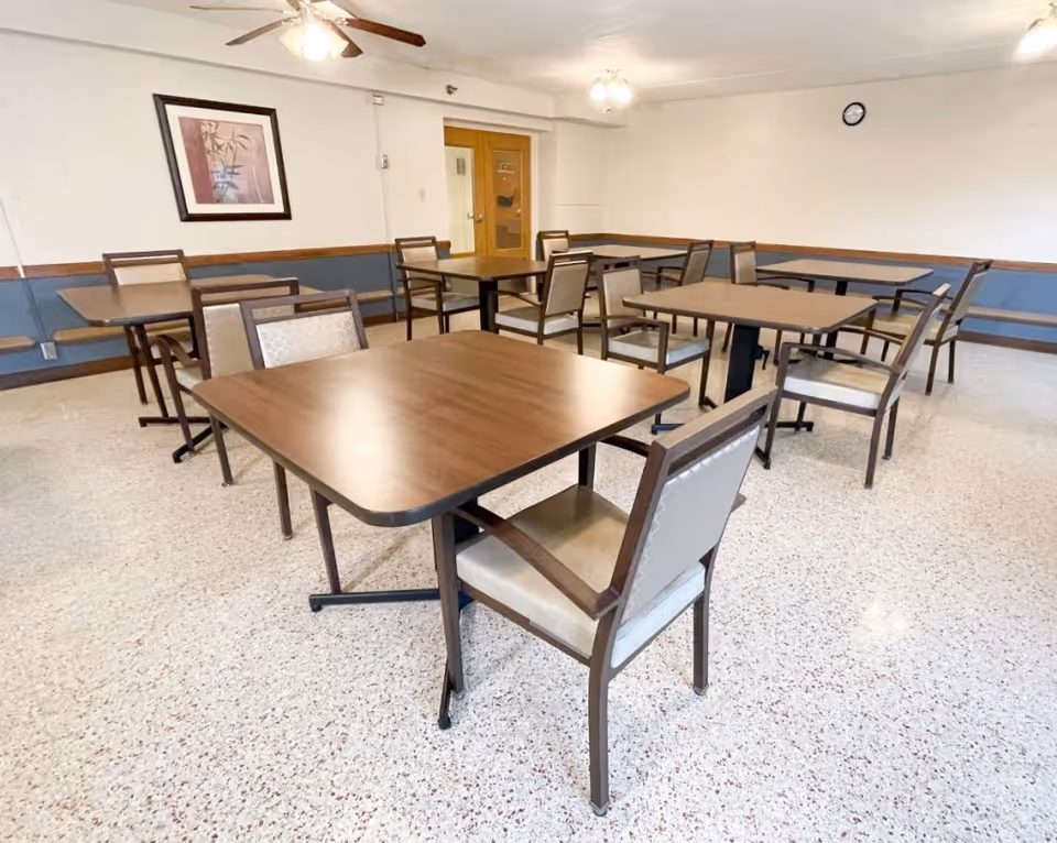 Community dining room with multiple wooden tables and cushioned chairs on a speckled floor under ceiling fans.
