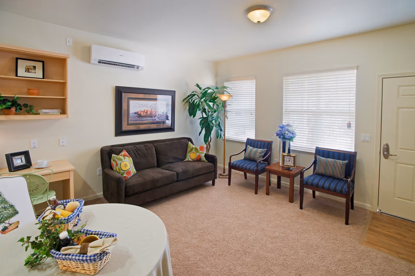 A cozy living room with a dark brown sofa adorned with colorful pillows, two blue striped armchairs with striped cushions, a small wooden side table with a framed photo and a vase of blue flowers, a tall floor lamp, and two windows with white blinds. There is a round table with a white tablecloth and a basket of fruit and plants in the foreground. A wooden desk with a green chair and some decorative items is on the left side, and a framed picture hangs on the wall above the sofa. The room has beige carpet and light-colored walls.