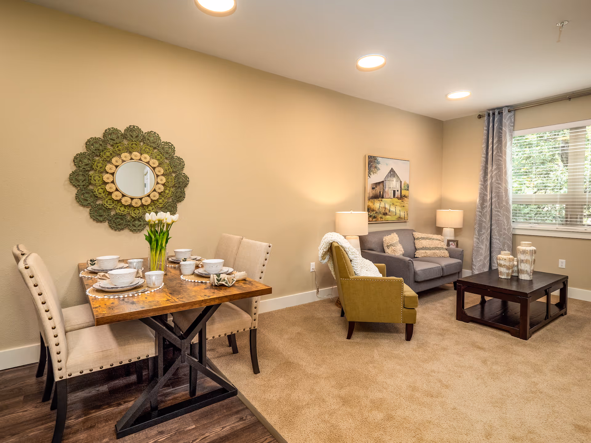 A cozy living and dining area in a senior living community. The dining area features a wooden table set with white dishes, cups, and a vase of white tulips, surrounded by four beige upholstered chairs. The living area has a gray loveseat with patterned pillows, a green armchair with a white throw, a dark wooden coffee table with decorative vases, two table lamps, a window with gray curtains, and a painting of a barn on the wall.