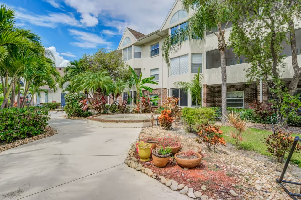 Landscaped courtyard with a paved walkway, potted plants, palm trees and a fountain beside a multi-story senior living building.