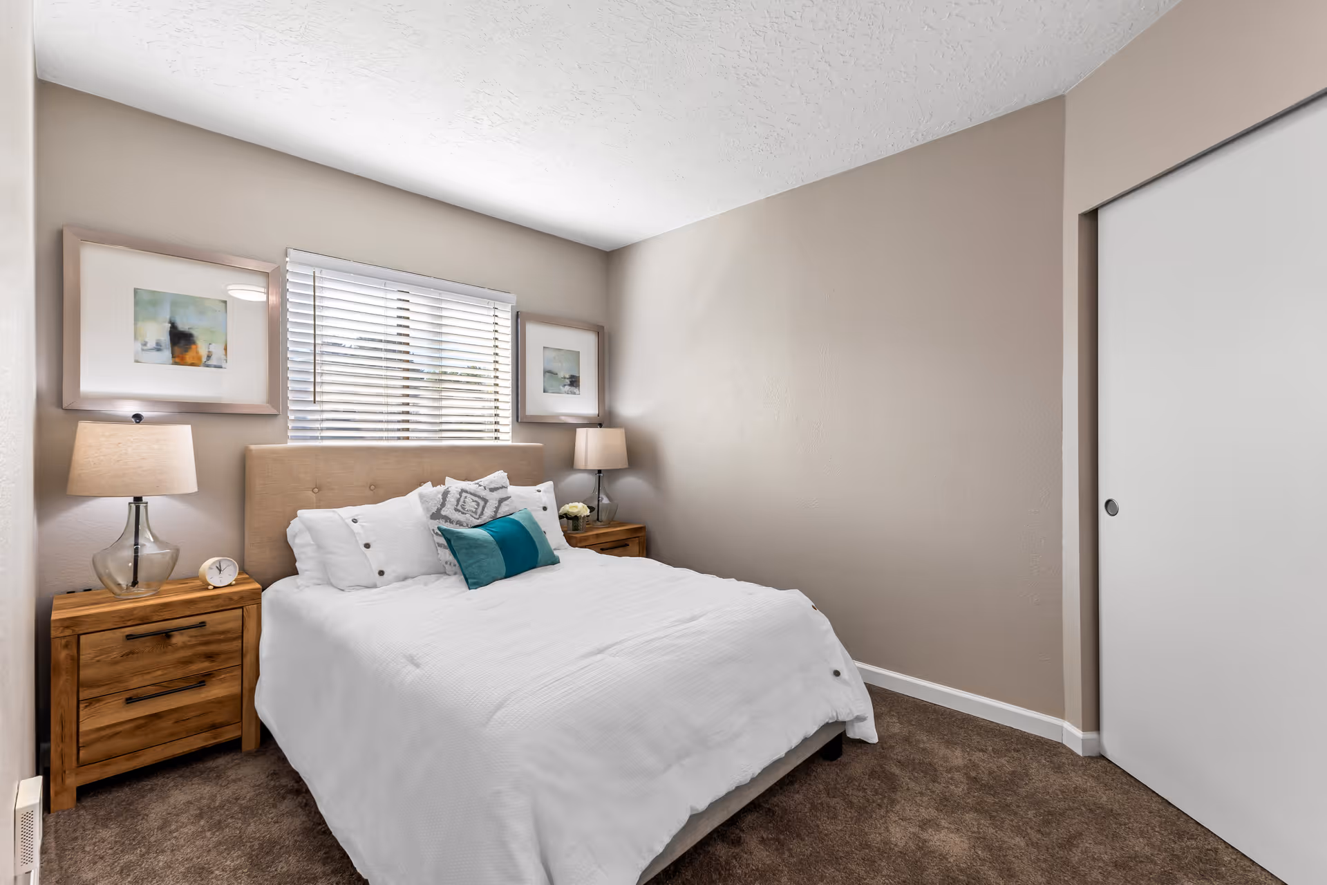 A cozy bedroom with a neatly made bed featuring white bedding and decorative pillows. The bed has a beige upholstered headboard and is flanked by two wooden nightstands, each with a lamp. There are two framed abstract paintings on the wall above the nightstands, and a window with white blinds is centered above the bed. The room has beige walls and brown carpet flooring, with a white sliding closet door on the right side.