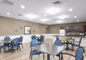 A spacious dining room with several square tables and blue cushioned chairs. The room has light-colored walls with decorative art pieces, wood flooring, and a kitchen area with dark wood cabinets and stainless steel appliances in the background. Ceiling lights provide bright illumination.
