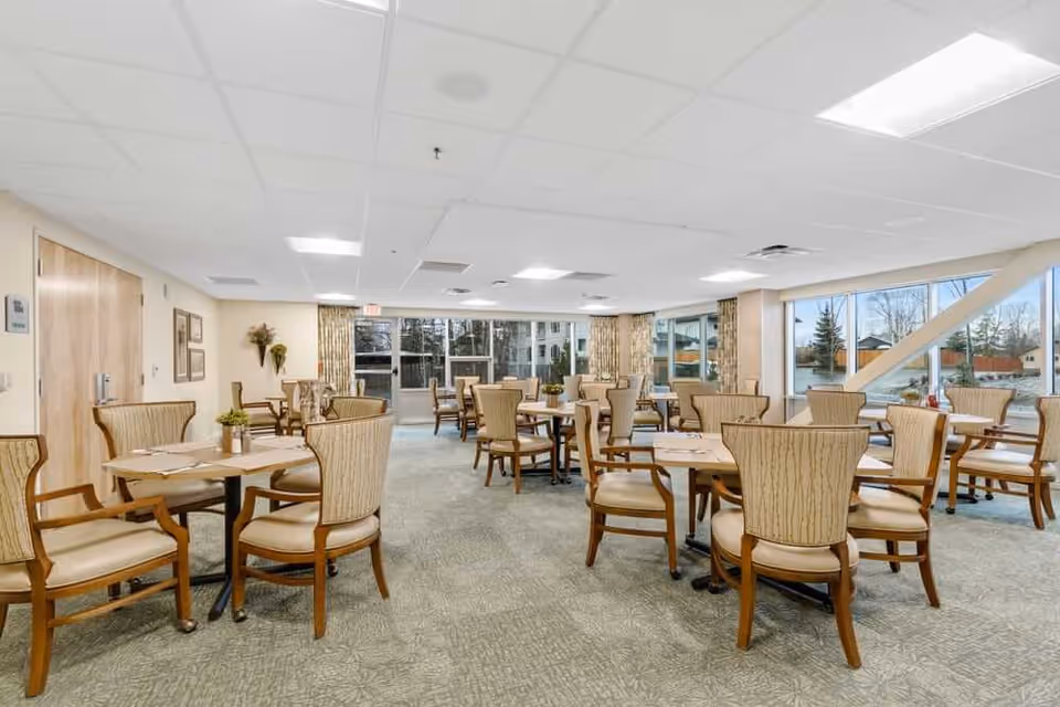 Bright senior living dining room with multiple round tables and upholstered chairs arranged near large windows.