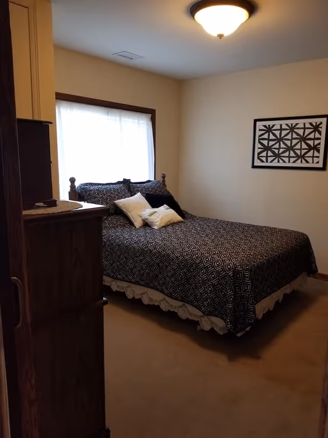 A bedroom with a bed covered in a black and white geometric patterned bedspread and several pillows. There is a wooden dresser to the left, a window with sheer white curtains behind the bed, and a framed black and white geometric artwork on the wall to the right. The room has beige walls and carpet, and a ceiling light fixture.