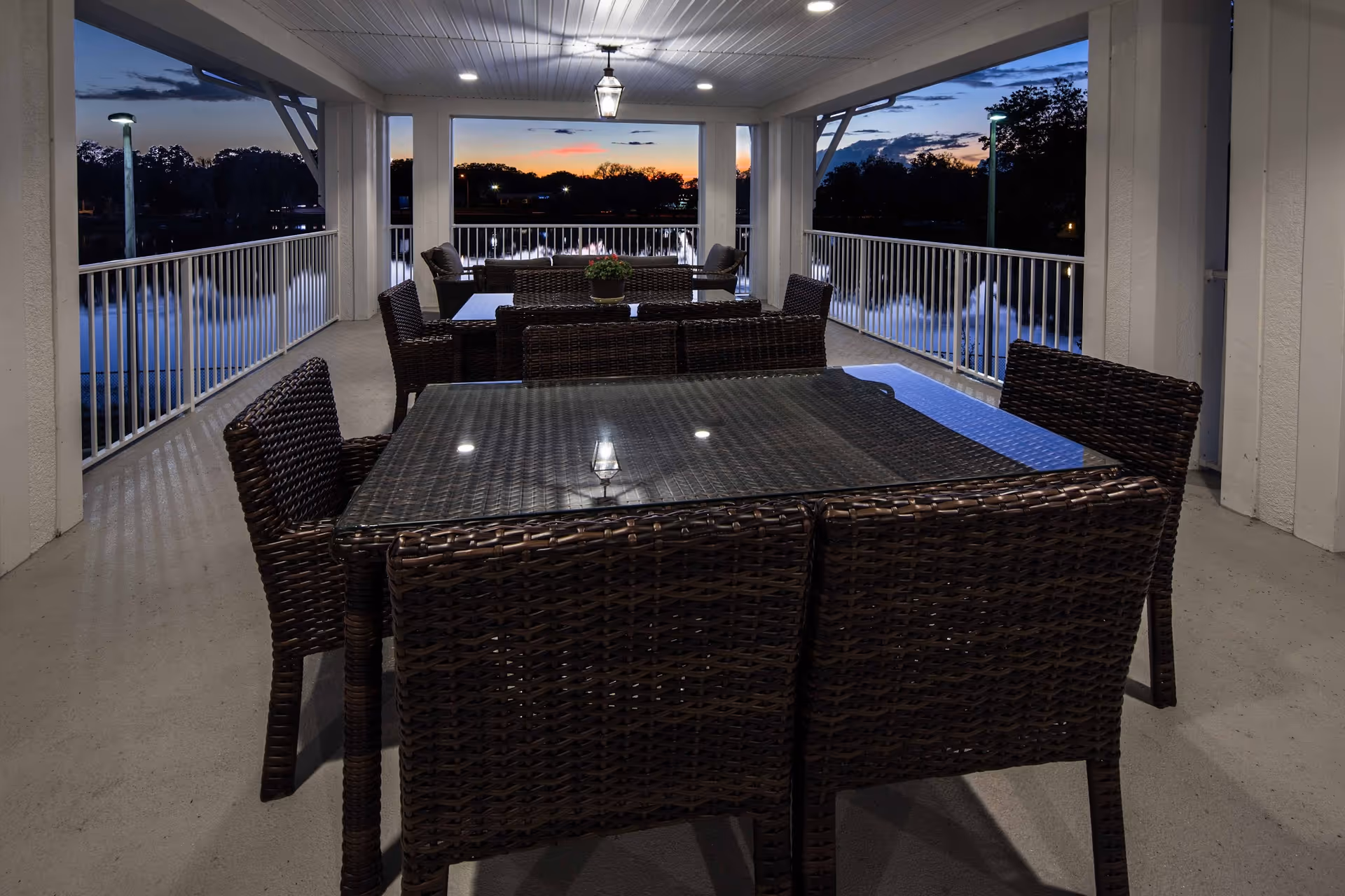 Covered outdoor patio area at dusk with multiple dark wicker tables and chairs. The patio overlooks a body of water with fountains and trees in the background under a colorful sunset sky.