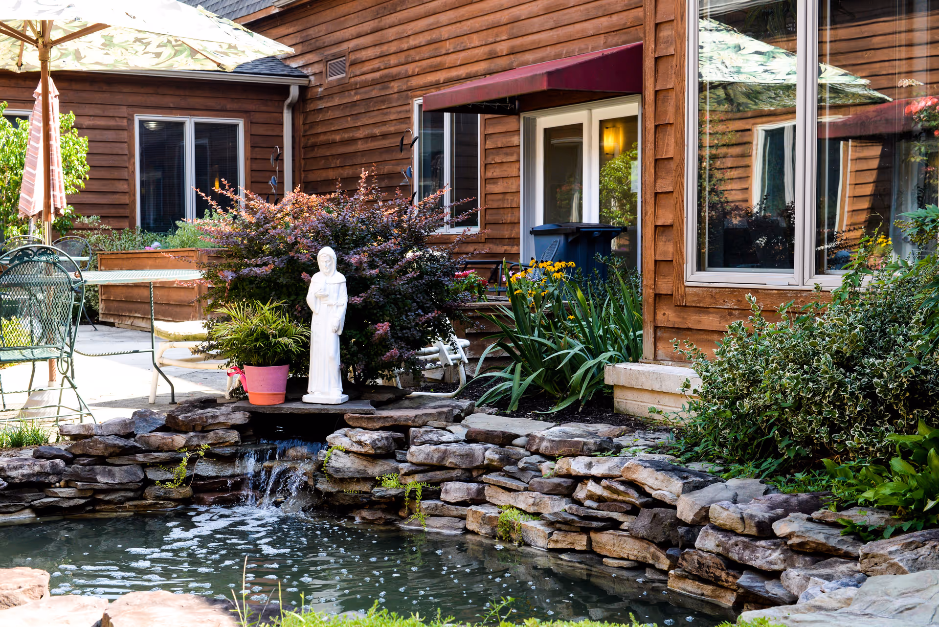 Outdoor courtyard area at Winter Growth Ruth Keeton House featuring a small pond with a stone border and a small waterfall. A white statue stands near the pond surrounded by plants and flowers. There are patio tables and chairs with umbrellas, and the building exterior is wooden with windows and a door with a maroon awning.