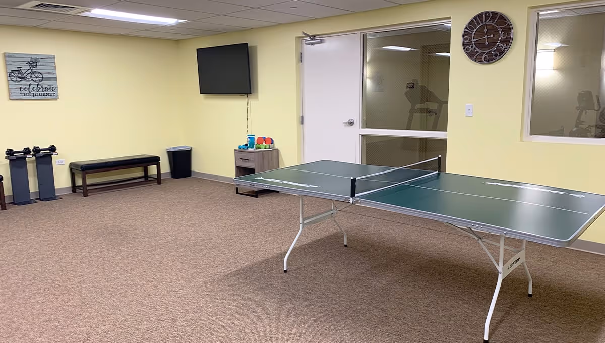 A recreational room with a ping pong table in the center, a wall-mounted TV, a bench, dumbbells on stands, a small cabinet with exercise balls and weights, a wall clock, and a motivational wall art that says 'celebrate the journey'. The walls are painted light yellow and the floor is carpeted.