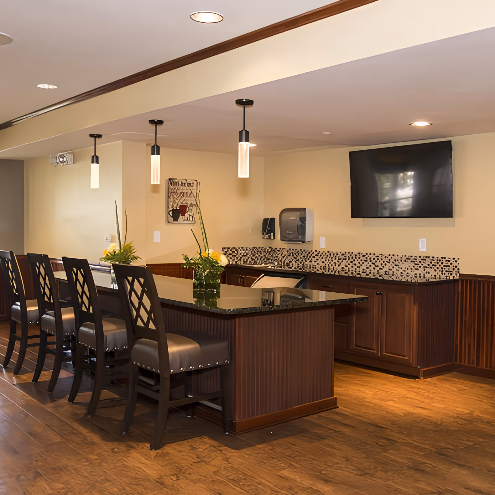 Interior view of a senior living facility's common area featuring a long counter with six high-back chairs. The counter has a dark granite top and wooden base, with two small flower arrangements placed on it. Above the counter, three pendant lights hang from the ceiling. On the wall behind the counter, there is a mounted flat-screen TV, a paper towel dispenser, and a soap dispenser. The floor is wooden, and the walls are painted a light beige color.