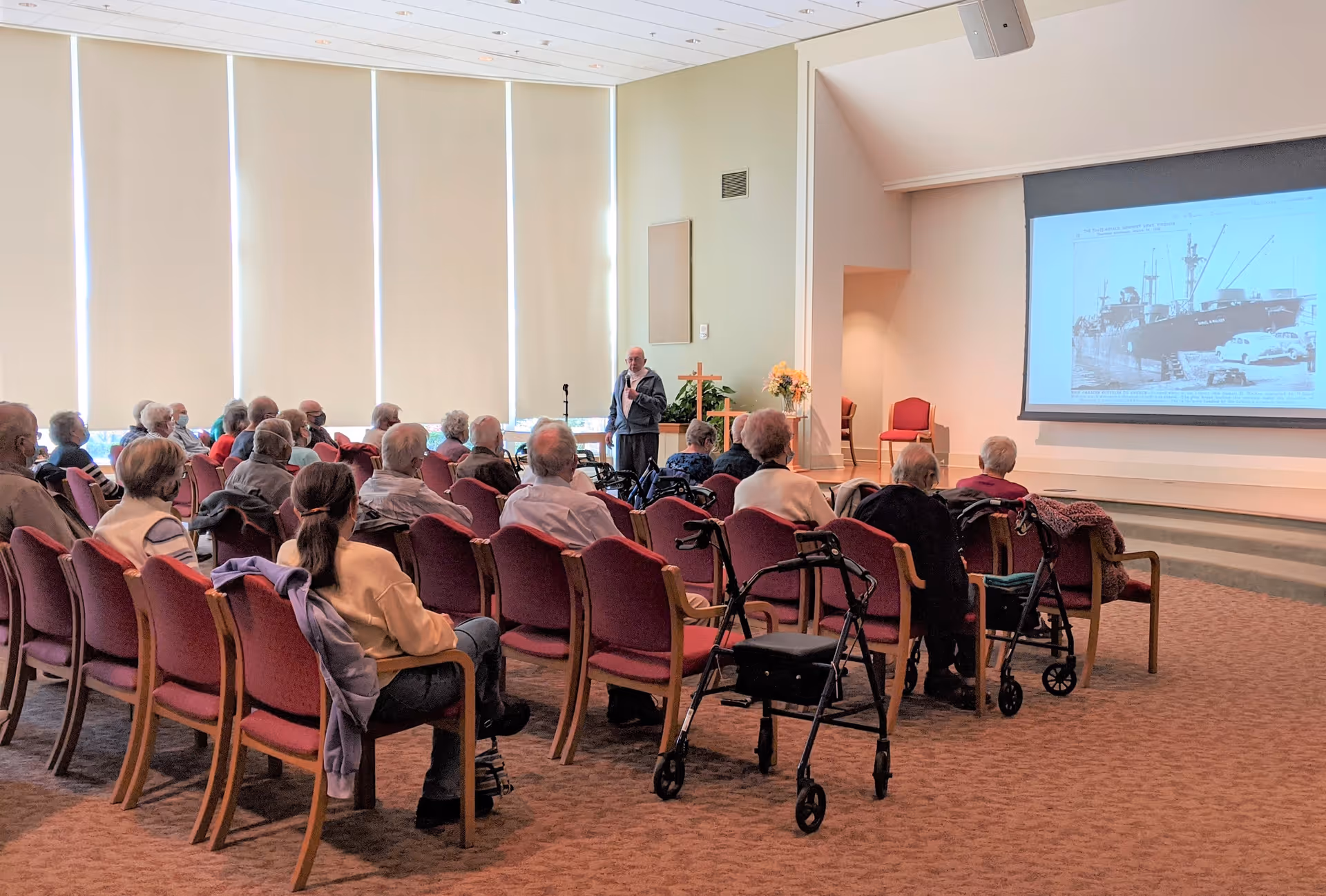 A group of elderly people seated in rows of chairs in a large room, attentively watching a presentation. A man stands near a microphone at the front of the room, speaking. There is a large projection screen displaying a black and white image of a ship and cars. Several walkers are visible among the seated audience. The room has tall windows with blinds and a carpeted floor.