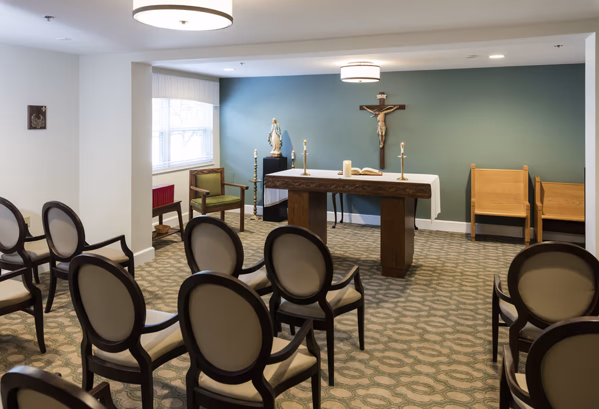Small chapel room with rows of chairs facing an altar topped with candles, a crucifix on a teal wall and religious statues.