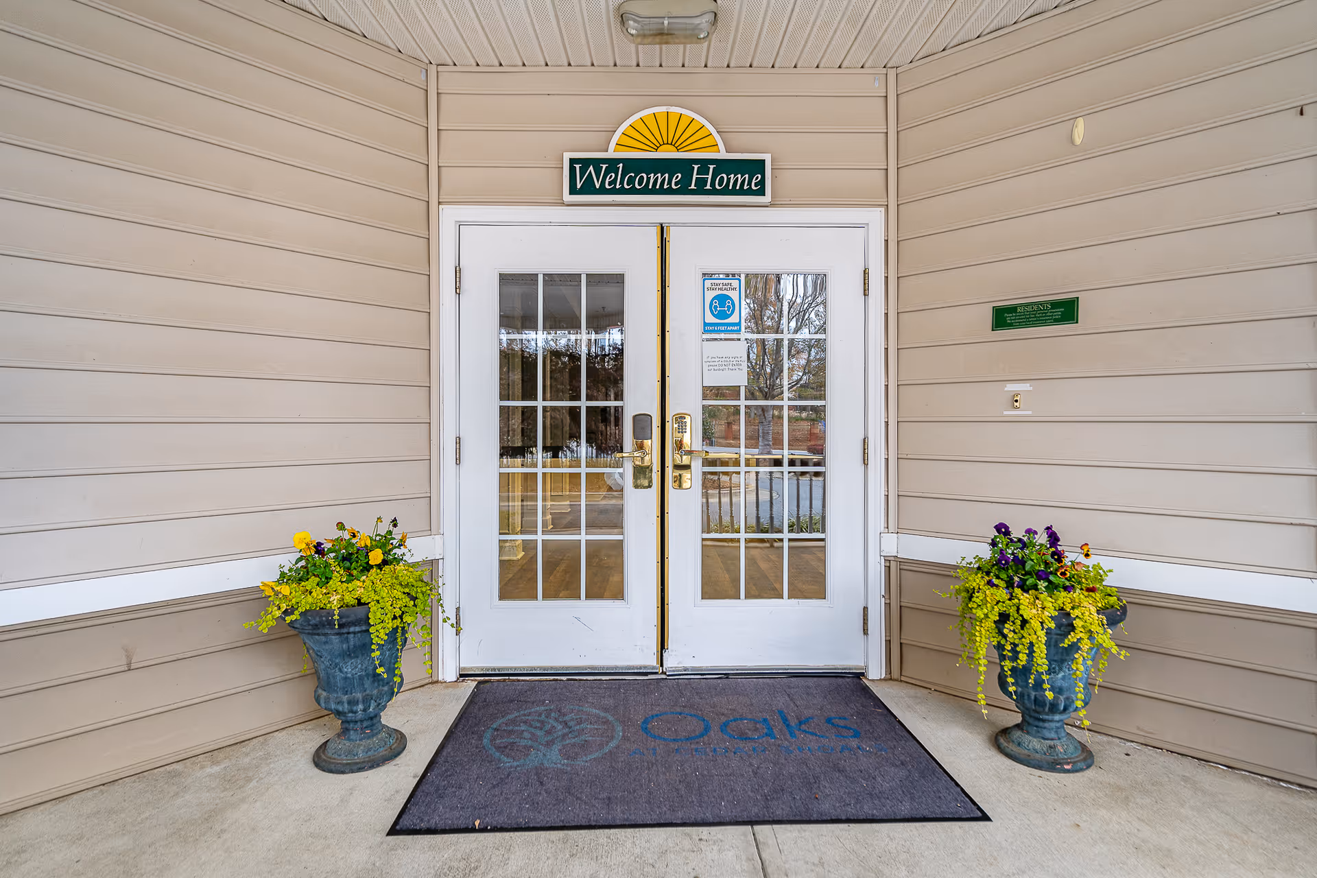 Entrance to a facility with double white doors featuring glass panes. Above the doors is a sign that reads 'Welcome Home'. On the ground in front of the doors is a mat with the logo and name 'Oaks at Cedar Shoals'. There are two large planters with yellow and purple flowers on either side of the entrance.