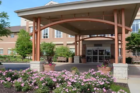 Covered porte-cochere entrance with glass doors on a brick building, framed by planters, benches, and pink flowering beds.