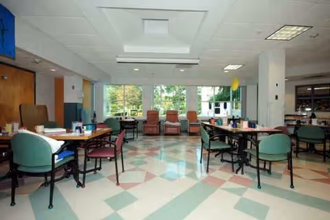 Bright common area with round tables, chairs, and large windows in a senior facility.