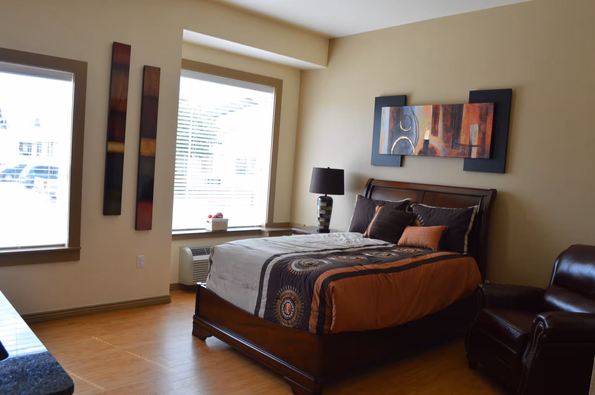 A bedroom in a senior living community featuring a wooden bed with a patterned brown and orange comforter and pillows. There is a dark brown leather armchair to the right of the bed, a bedside table with a lamp, and two large windows with blinds letting in natural light. The walls are beige with modern abstract artwork hanging above the bed and beside the window.