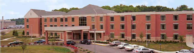 Exterior view of a large, three-story brick building with multiple windows and a covered entrance. Several cars are parked in front of the building, and there is a grassy area with small trees and shrubs surrounding the driveway.