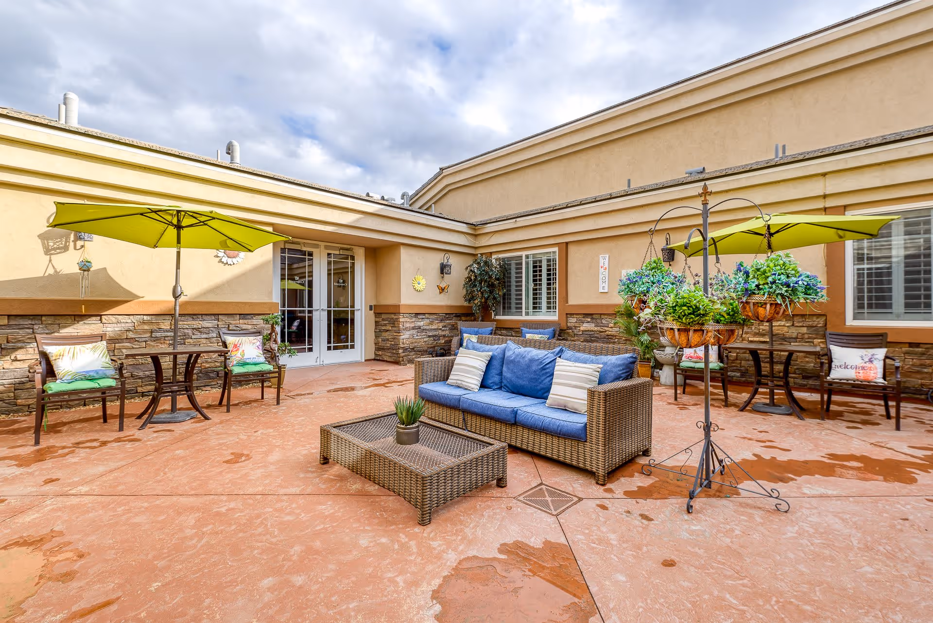 Outdoor patio area at San Martin Senior Living with wicker sofa and coffee table, green umbrellas shading wooden benches with colorful cushions, potted plants, and a beige building exterior under a partly cloudy sky.