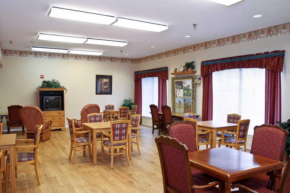 Spacious common dining room with several wooden tables and upholstered chairs, large windows with red curtains, and a TV cabinet.