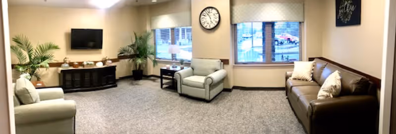 A cozy living room area with two armchairs and a brown leather sofa, all with cushions. There is a wall-mounted flat-screen TV above a dark wooden cabinet, two large potted plants, a clock on the wall, and two windows with blinds letting in natural light.
