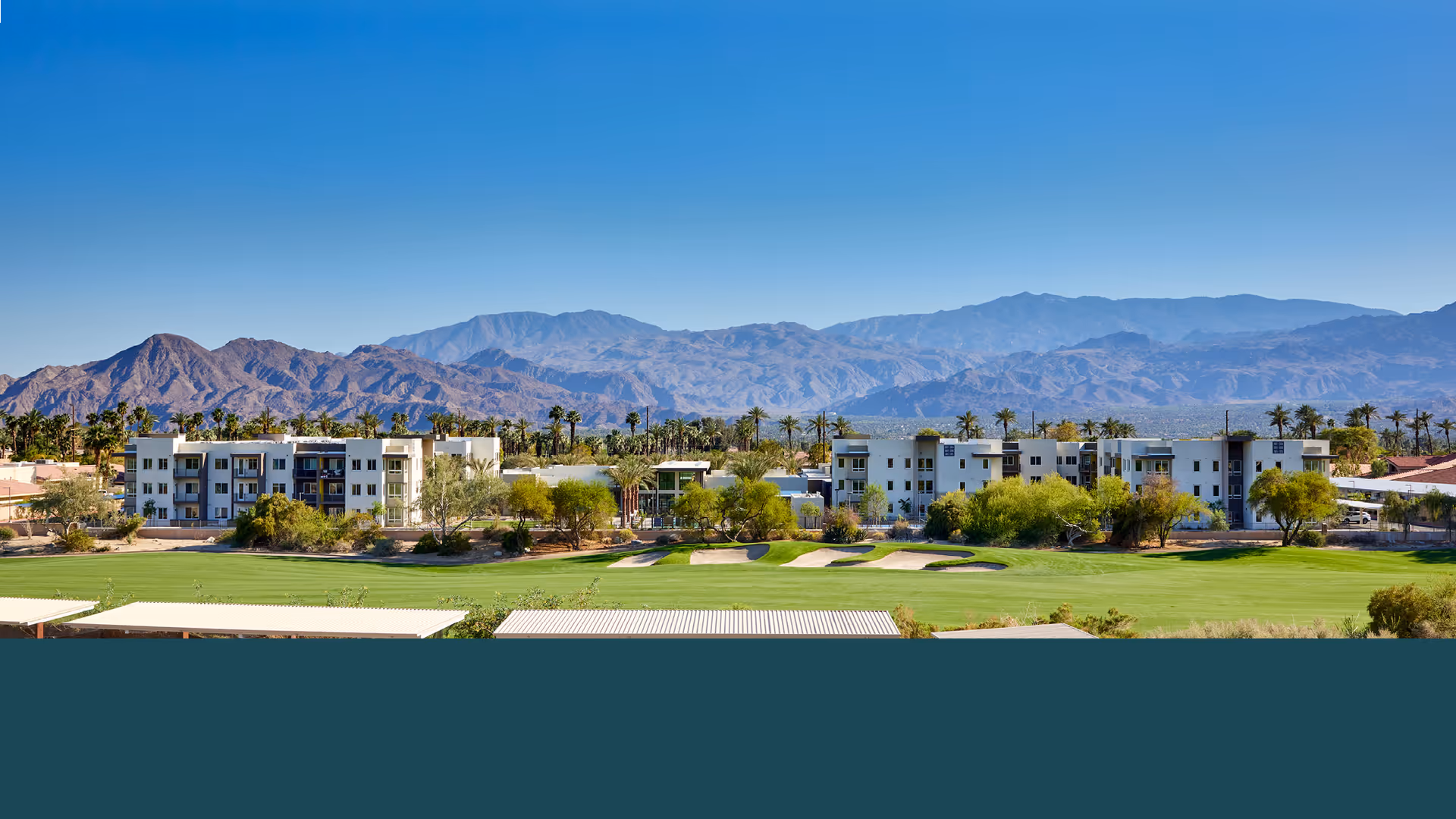 Low-rise residential buildings by a golf course with palm trees and mountains in the background under a clear blue sky.