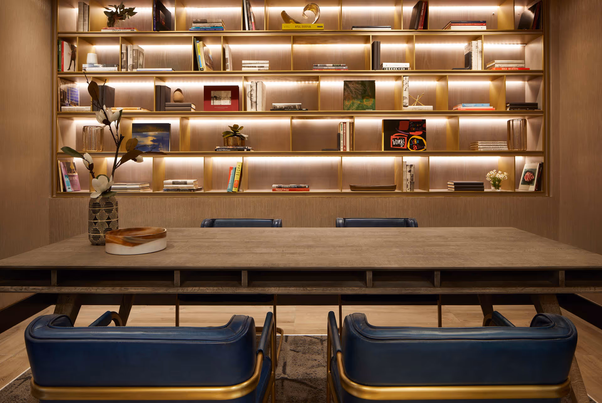 A modern interior room featuring a large wooden table with four blue leather chairs around it. Behind the table is a wall-mounted bookshelf with built-in lighting, displaying various books, decorative items, and plants. A vase with flowers and a decorative bowl are placed on the table.