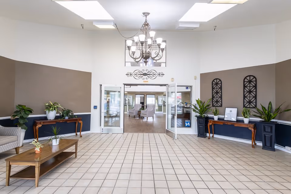 A spacious and well-lit lobby area with tiled flooring, a chandelier hanging from the ceiling, and two wooden tables adorned with potted plants on either side. There are two open glass doors leading to a room with armchairs and large windows in the background.