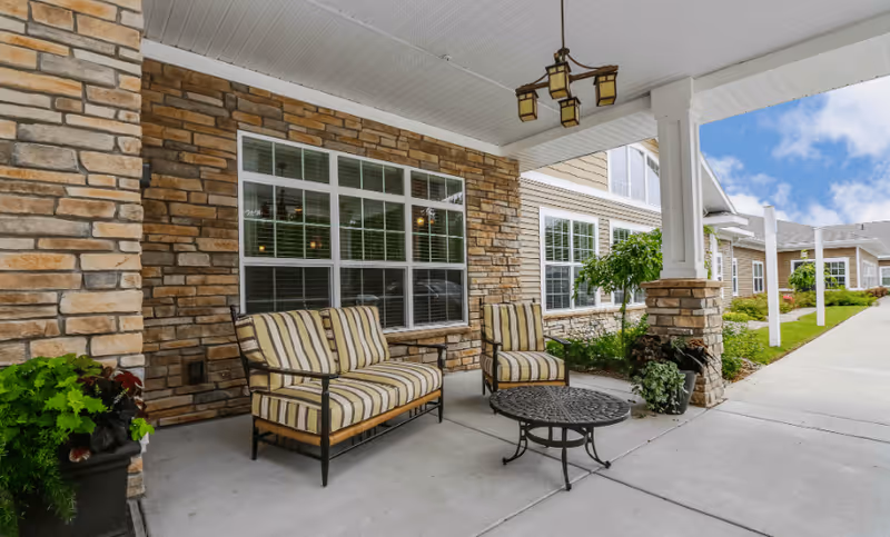 Covered outdoor seating area at Candlestone Assisted Living & Memory Care with striped cushioned chairs and a small round metal table, stone wall exterior, large windows, potted plants, and a hanging light fixture under a white ceiling.