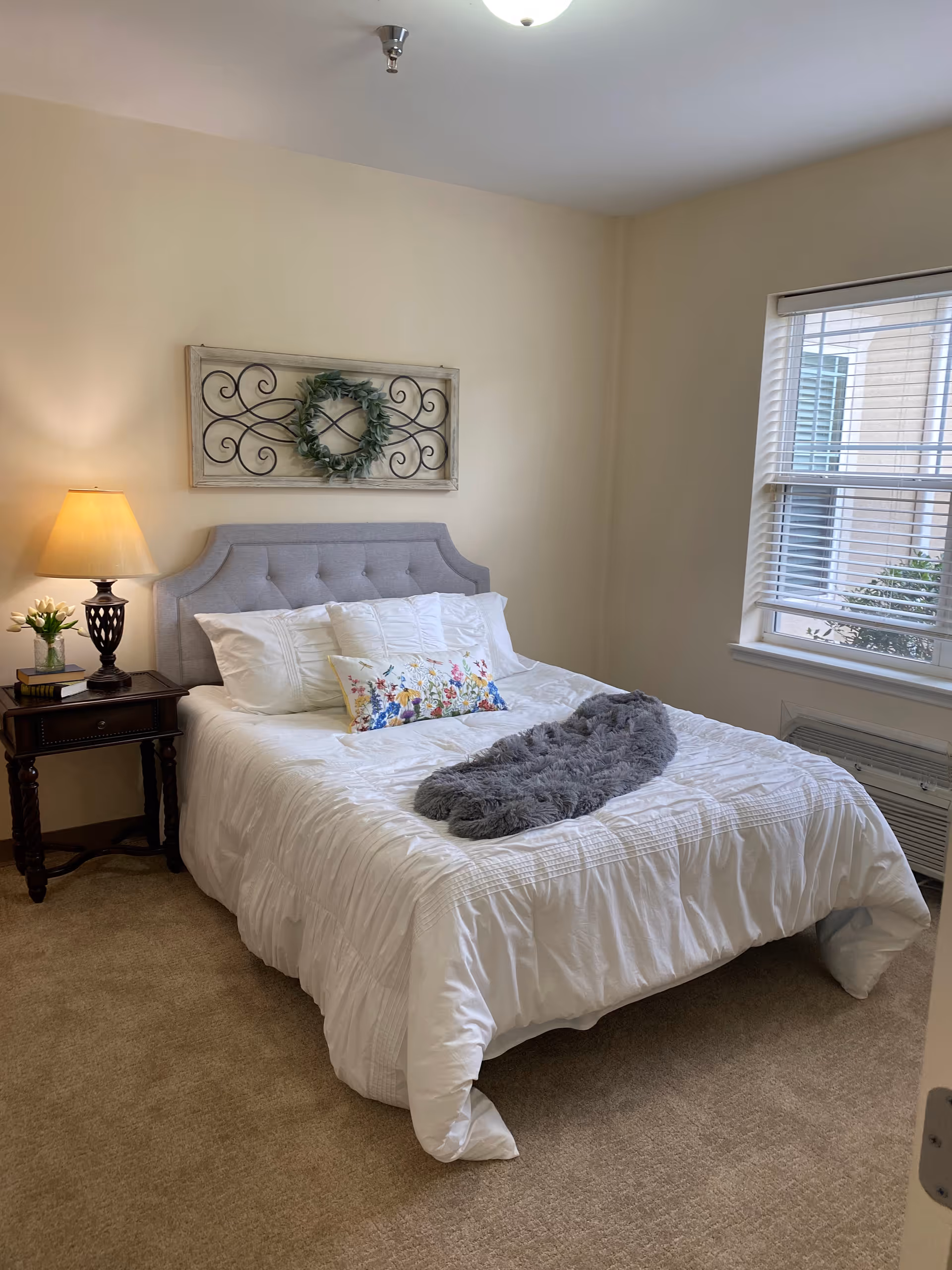 A furnished bedroom with a tufted headboard, white bedding, a nightstand and lamp, wall decor, and a window.