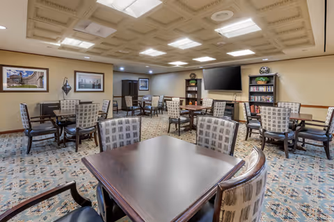 A spacious common area with multiple square wooden tables and cushioned chairs arranged neatly on a patterned carpet. The room has a decorative ceiling with recessed lighting and framed pictures on the walls. There is a large flat-screen TV mounted on the wall above bookshelves filled with books and decorative items.