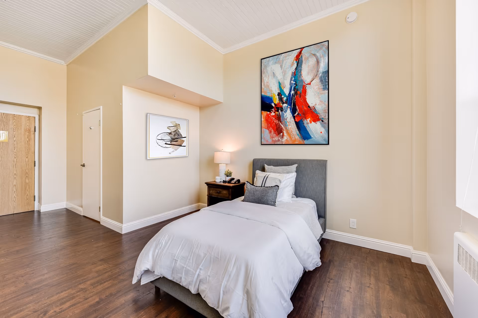 A neatly made single bed with white bedding and multiple pillows in a room with beige walls and wooden flooring. There is a wooden nightstand with a lamp, a phone, and a small plant next to the bed. Two abstract paintings hang on the walls, one above the bed and one on the adjacent wall. A wooden door and a small closet door are visible in the background.
