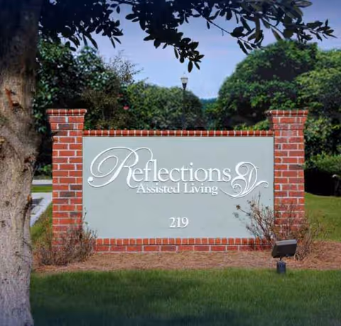 Brick entry sign reads Reflections Assisted Living with landscaping and trees in the background.