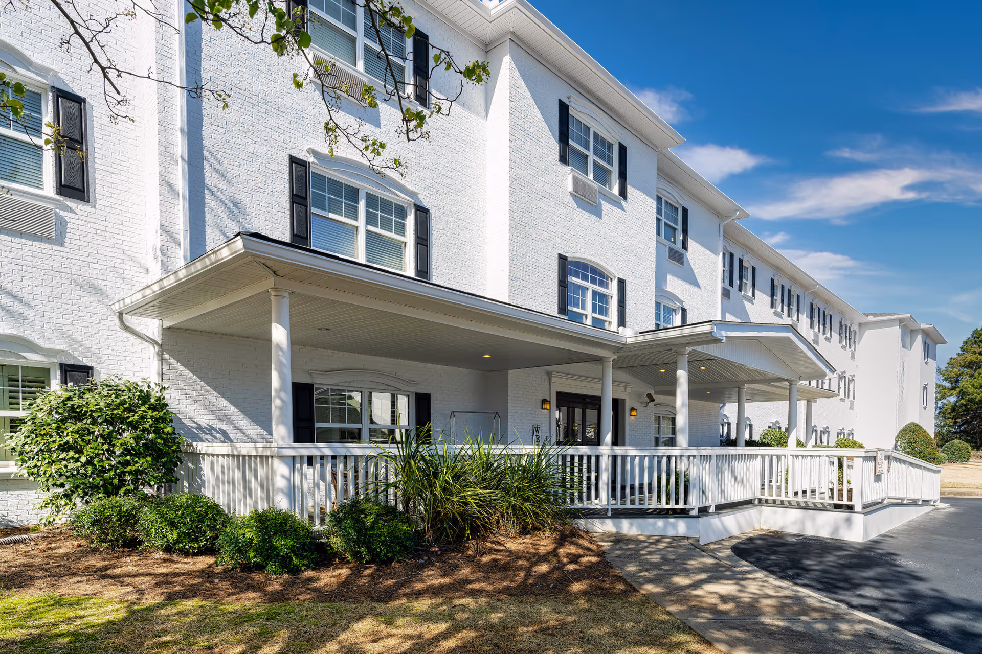 Exterior view of a white brick senior living facility building with black window shutters, a covered entrance with white columns, a wheelchair accessible ramp, and landscaped bushes under a clear blue sky.