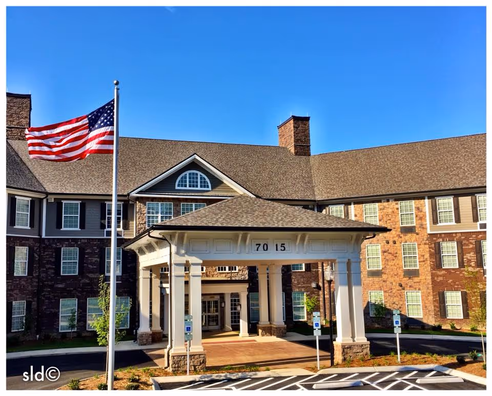 Front exterior view of a senior living facility building with a covered entrance, multiple windows, and an American flag on a flagpole. The building has a brick and siding facade under a clear blue sky.